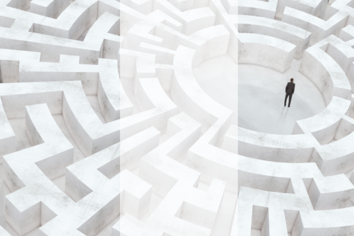 A person in formal attire stands at the centre of a large, white, three-dimensional maze. The maze walls are tall and intricate, symbolising complexity and challenges. The scene conveys a sense of isolation and problem-solving within a vast, structured environment.